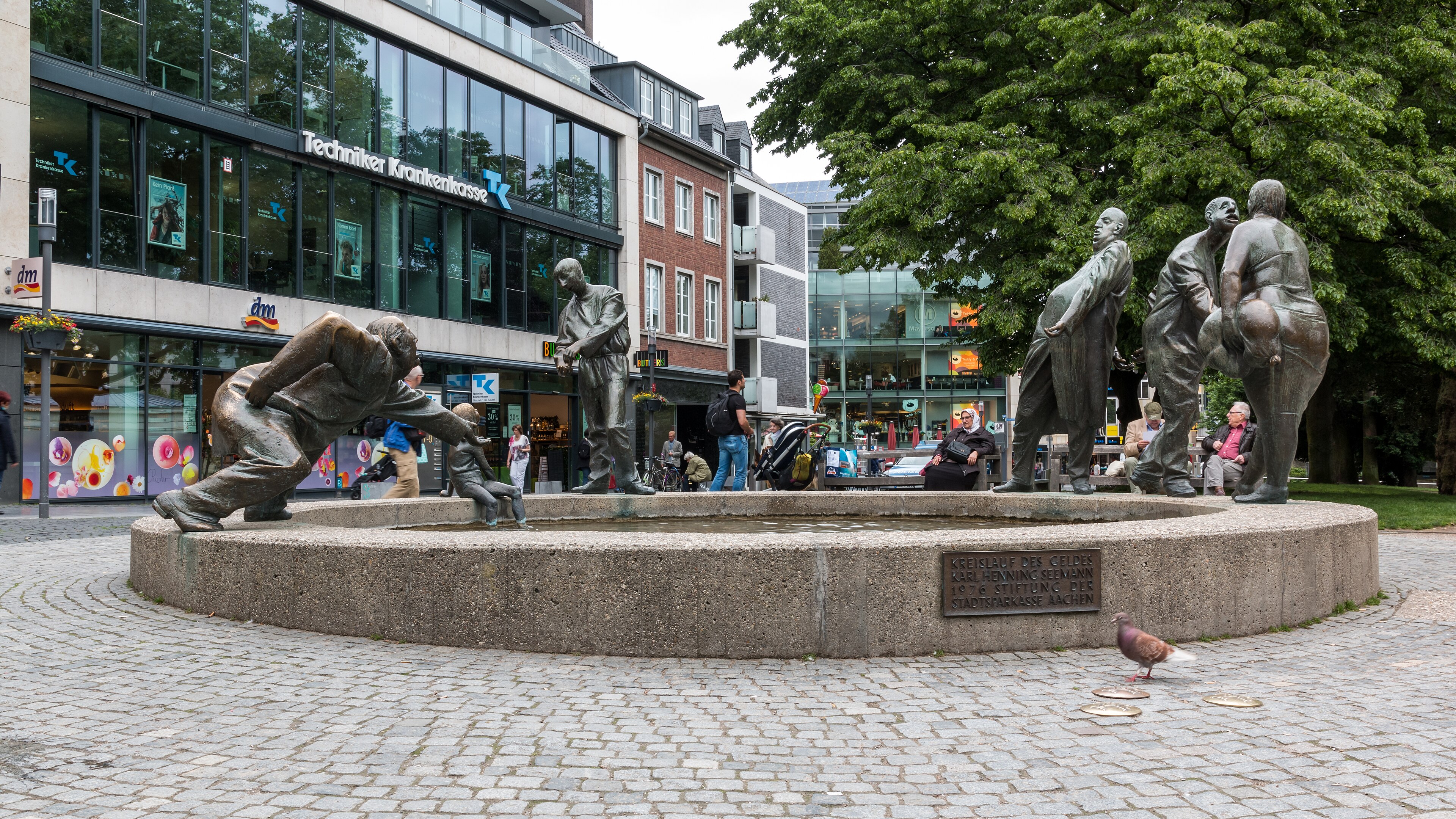 Brunnen „Kreislauf des Geldes“ in Aachen mit mehreren Bronzefiguren um ein rundes Wasserbecken